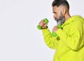 Middle aged serious brutal bearded man in bright yellow green casual hoodie standing and holding green dumbbels in hands