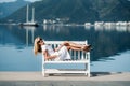 Middle aged sexy smiling caucasian woman with sunglasses sits on sea shore in white dress