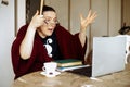 Middle-aged surprised woman wearing vinous cardigan, glasses sitting at table at home, raising hands, looking at laptop. Royalty Free Stock Photo