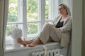 A middle-aged woman in a beige suit and black tank top sits mysteriously and looks out the window on the windowsill Royalty Free Stock Photo