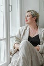 A middle-aged woman in a beige suit and black tank top sits mysteriously and looks out the window on the windowsill Royalty Free Stock Photo