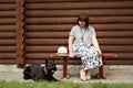 Middle-aged woman in a countryside sitting on a wooden bench and looking at a black schnauzer near a log house