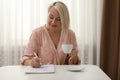 Middle aged woman with cup of drink solving puzzle at table indoors Royalty Free Stock Photo