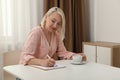 Middle aged woman with cup of drink solving puzzle at table indoors Royalty Free Stock Photo