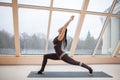 Middle aged woman doing yoga in Virabhadrasana One or Warrior One yoga pose on the mat in front of large windows., exercise fitnes Royalty Free Stock Photo