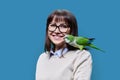 Middle aged woman with green Quaker parrot on her shoulder, on blue background