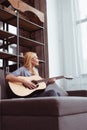 middle aged woman playing guitar and looking away while sitting on sofa Royalty Free Stock Photo