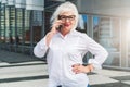 A middle-aged woman, a retired woman dressed in a white shirt and glasses, stands on a city street and talks on phone Royalty Free Stock Photo