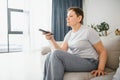 middle-aged woman sitting on the sofa and turning on the TV with a remote control. Royalty Free Stock Photo