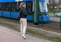 Middle-aged woman of a sports type in a jacket and sweatpants stands at a tram stop next to a tram, urban spring landscape