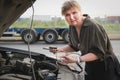 Middle-aged woman stands in front of a car with the hood open and holds the battery charging clamps