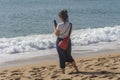 A middle-aged WOMAN stands on a sandy beach and takes pictures of the sea on her smartphone, back view. Royalty Free Stock Photo