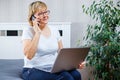 A middle-aged woman talking on the phone and working on laptop at home. Royalty Free Stock Photo