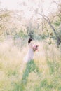 In the middle of the sunny park the bride in long white dress is holding the huge pink bouquet. Back view.