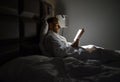 Midle-aged Caucasian man relaxing in bed reading bestseller novel paper book with bedside lamp turned on. Evening relaxation,