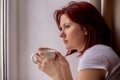 Middle-aged woman thoughtfully looking at the window, holding in hands white mug of tea or coffee. Short break from work or chores Royalty Free Stock Photo