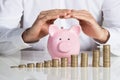 Businessman Protecting Piggybank With Stacked Coins At Desk