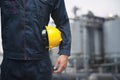 Midsection of young worker holding a yellow hardhat outdoors with factory in the background