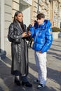 Woman with black leather trench coat and grey turtleneck and man with blue Moncler puffer jacket