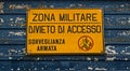 Military area with yellow sign: `Zona militare, divieto di accesso, sorveglianza armata`. Translation: `Milita