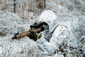 Military man in white camouflage uniform with hood and machinegun in the long winter grass. Soldier stood on knelt and aims of the