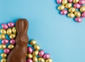 Milk chocolate Easter bunny and frame of pile of candy eggs wrapped in pink and golden foil on blue background