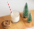 Milk for Santa. Glass milk, drinking straw, cookies, decorative Xmas trees on wooden board on white.