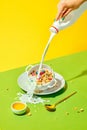 Milky breakfast. Woman pouring milk into cereal bowl, muesli with berries against green yellow background