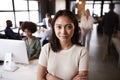 Millennial Asian female creative standing in a busy casual office, smiling to camera