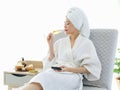 Millennial Asian female customer in white clean bathrobe and towel sitting on armchair drinking sparkling champagne while surfing Royalty Free Stock Photo