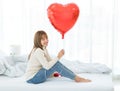 Millennial Asian young beautiful female teenager in turtleneck sweater and jeans sitting on bed smiling holding red heart shape Royalty Free Stock Photo