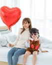 Millennial Asian young beautiful mother sitting on bed smiling holding red heart shaped air balloon while little cute preschooler Royalty Free Stock Photo