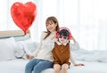 Millennial Asian young beautiful mother sitting on bed smiling holding red heart shaped air balloon while little cute preschooler Royalty Free Stock Photo