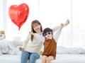 Millennial Asian young beautiful mother sitting on bed smiling holding red heart shaped air balloon while little cute preschooler Royalty Free Stock Photo