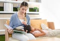 Millennial Asian young female mother sitting on sofa couch reading bedtime story book to little girl daughter laying lying down on Royalty Free Stock Photo