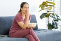 Millennial Asian young happy cheerful female housewife sitting smiling on cozy sofa smiling holding eating popcorn in glass bowl Royalty Free Stock Photo