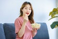 Millennial Asian young happy cheerful female housewife sitting smiling on cozy sofa smiling holding eating popcorn in glass bowl Royalty Free Stock Photo