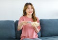 Millennial Asian young happy cheerful female housewife sitting smiling on cozy sofa holding popcorn snack glass bowl using remote Royalty Free Stock Photo