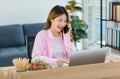 Millennial Asian young teenagers female freelancer worker sitting holding talking via smartphone while working with laptop Royalty Free Stock Photo