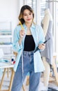 Millennial Asian young thougtful professional female dressmaker designer seamstress with measuring tape standing holding pencil