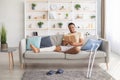 Millennial black guy with plastered leg sitting on sofa, reading book at home, pair of crutches standing nearby Royalty Free Stock Photo