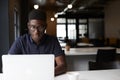 Millennial black male creative sitting at a desk in an open plan office using laptop, close up Royalty Free Stock Photo