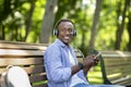 Millennial black man sitting on bench in park and listening to music from mobile phone Royalty Free Stock Photo