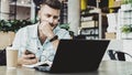 Millennial businessman sitting in cafe with open laptop, looking thoughtfully at computer screen, holding smartphone.