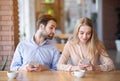 Millennial couple having dull date, stuck in smartphones, jealous young man looking into woman`s gadget at cafe