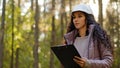 Millennial female technician ecologist looking up at treetops, Young indian woman in hardhat with clipboard taking