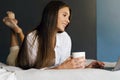 Millennial girl is resting on bed in white shirt, holding cup of coffee in her hand.