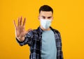 Millennial guy in face mask gesturing STOP on orange studio background