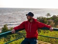 Millennial man in bucket hat red hoody eyeglasses on bench with autumn sea view. Authentic male tourist lifestyle photo