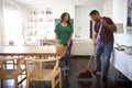 Millennial man sweeping the floor in the dining room while his partner stands talking to him